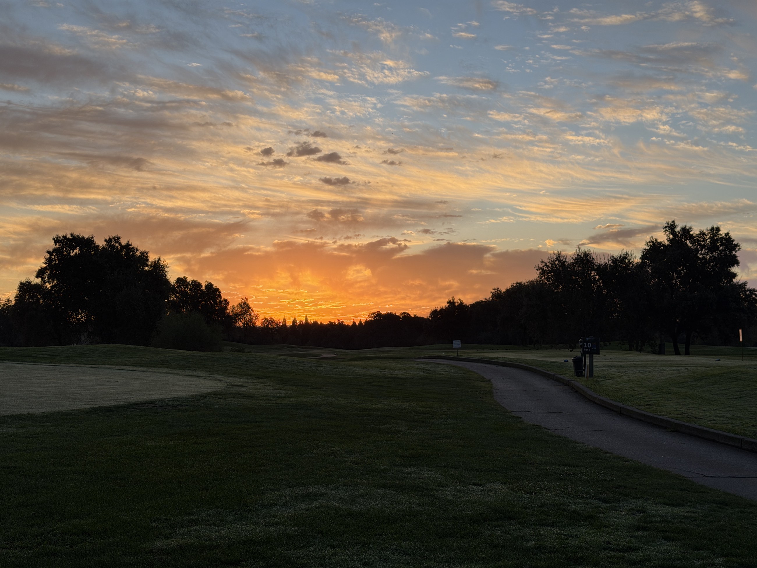 Sunrise over the tenth at Woodcreek Golf Club, valley oaks in silhouette against an orange sky, the cart path curving away to the right