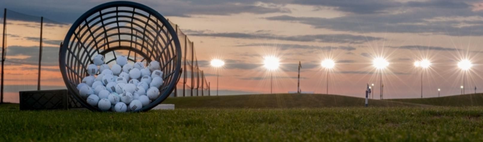 The lighted driving range at Woodcreek glowing at dusk, a basket of range balls in the foreground
